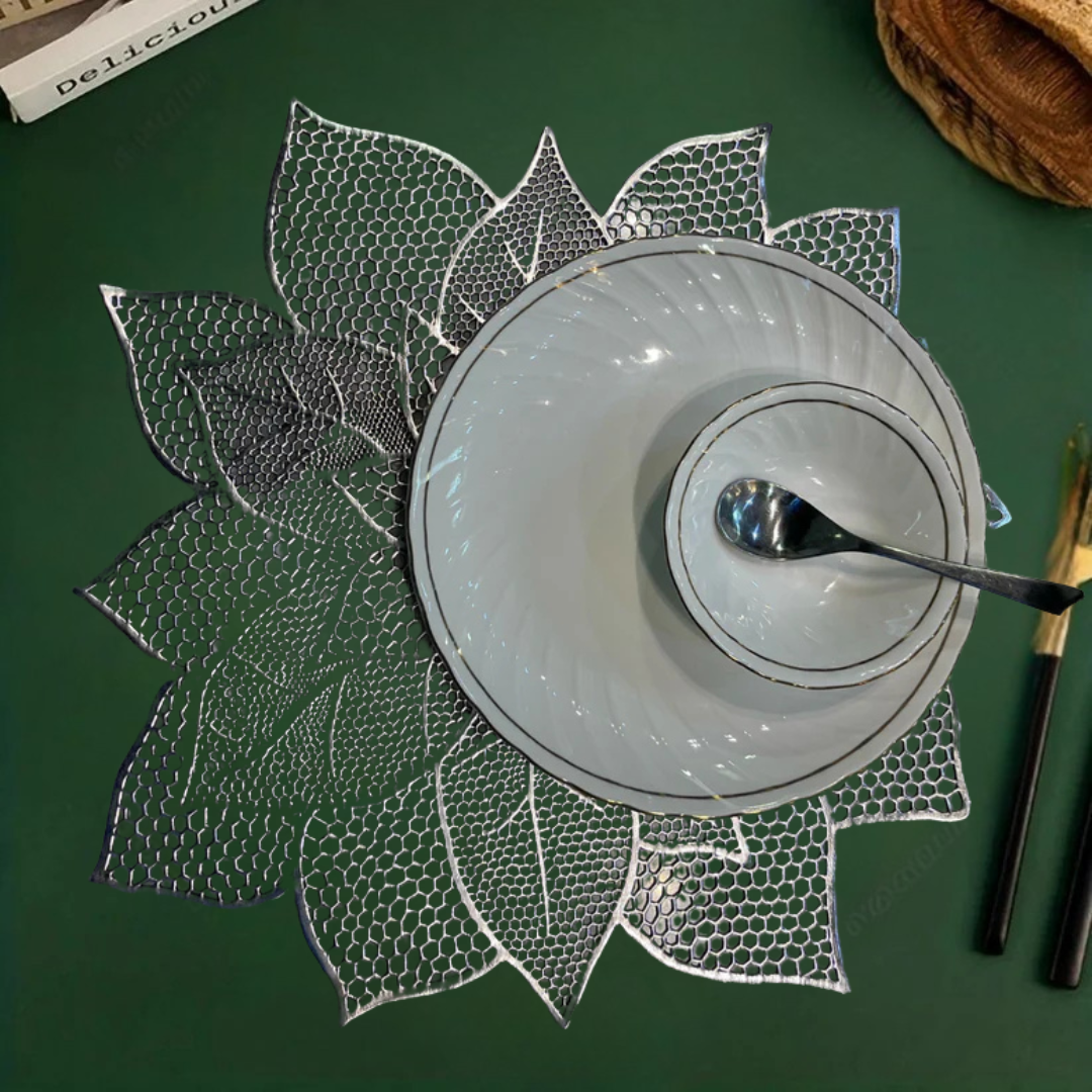 White bowl and small dish with spoon on a lace-like, leaf-shaped silver placemat over a green surface.