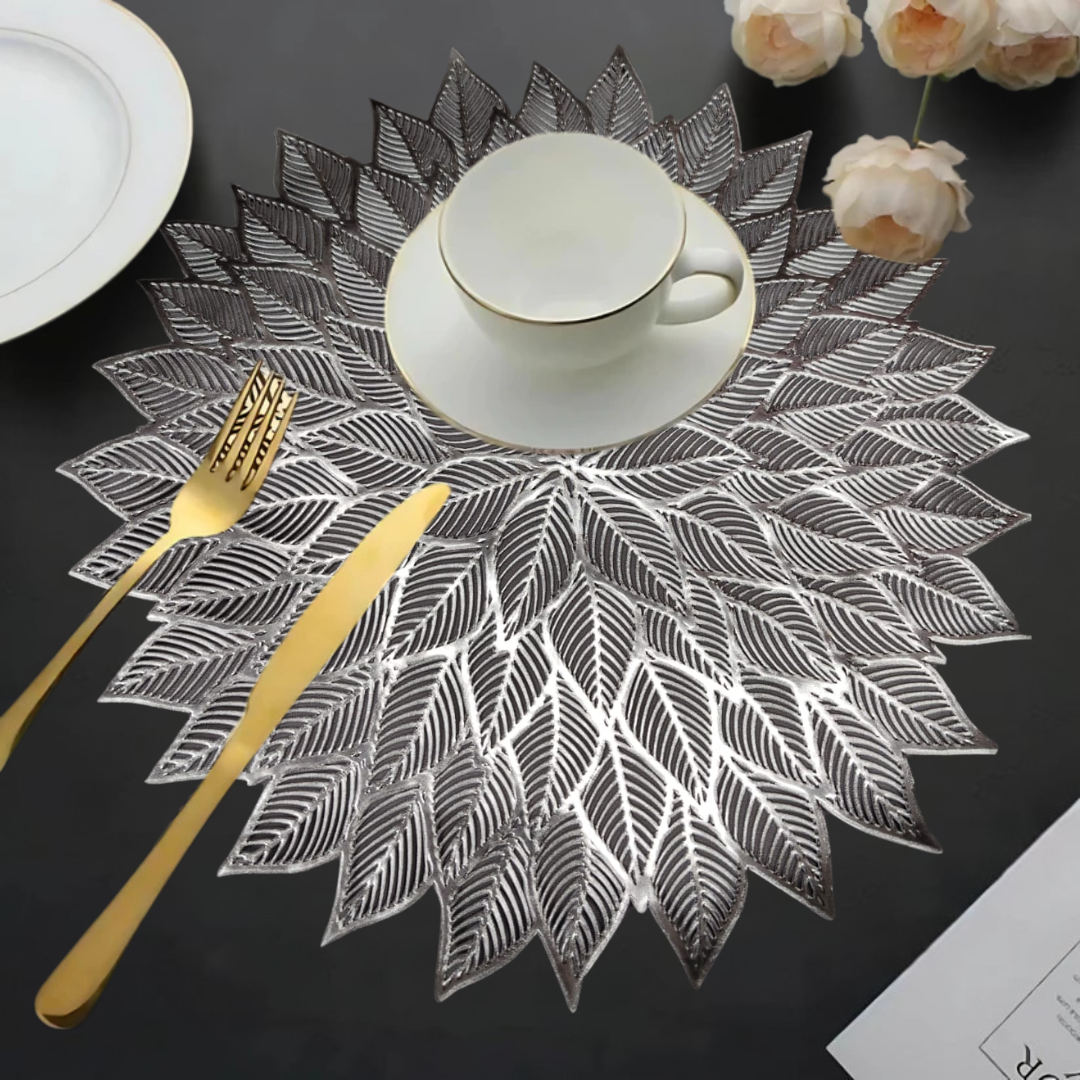 Decorative leaf-patterned silver placemat with a white cup, gold cutlery, and flowers on a dark table.