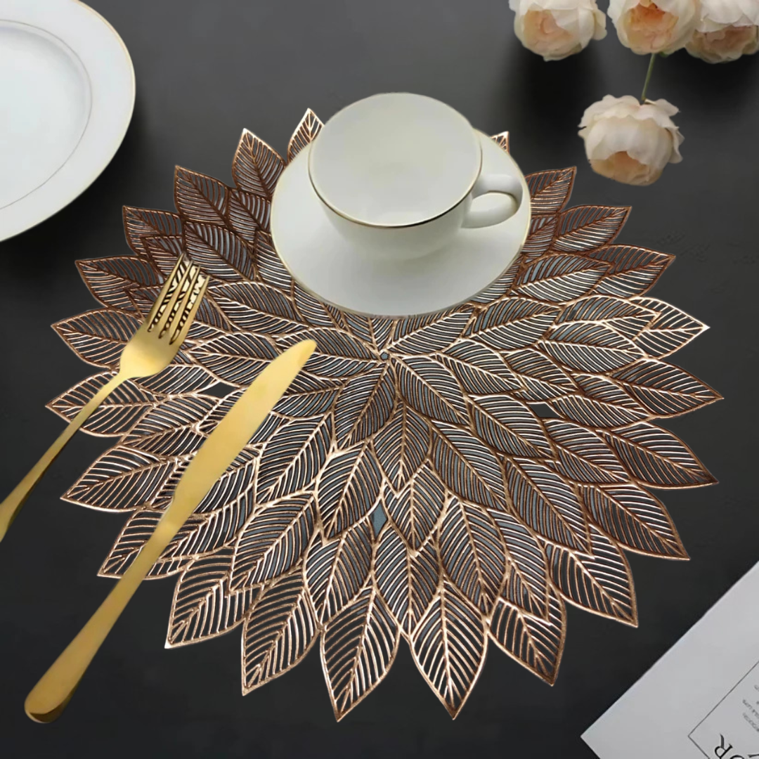 Decorative leaf-patterned copper table mats with a white cup, gold cutlery, and flowers on a dark table.