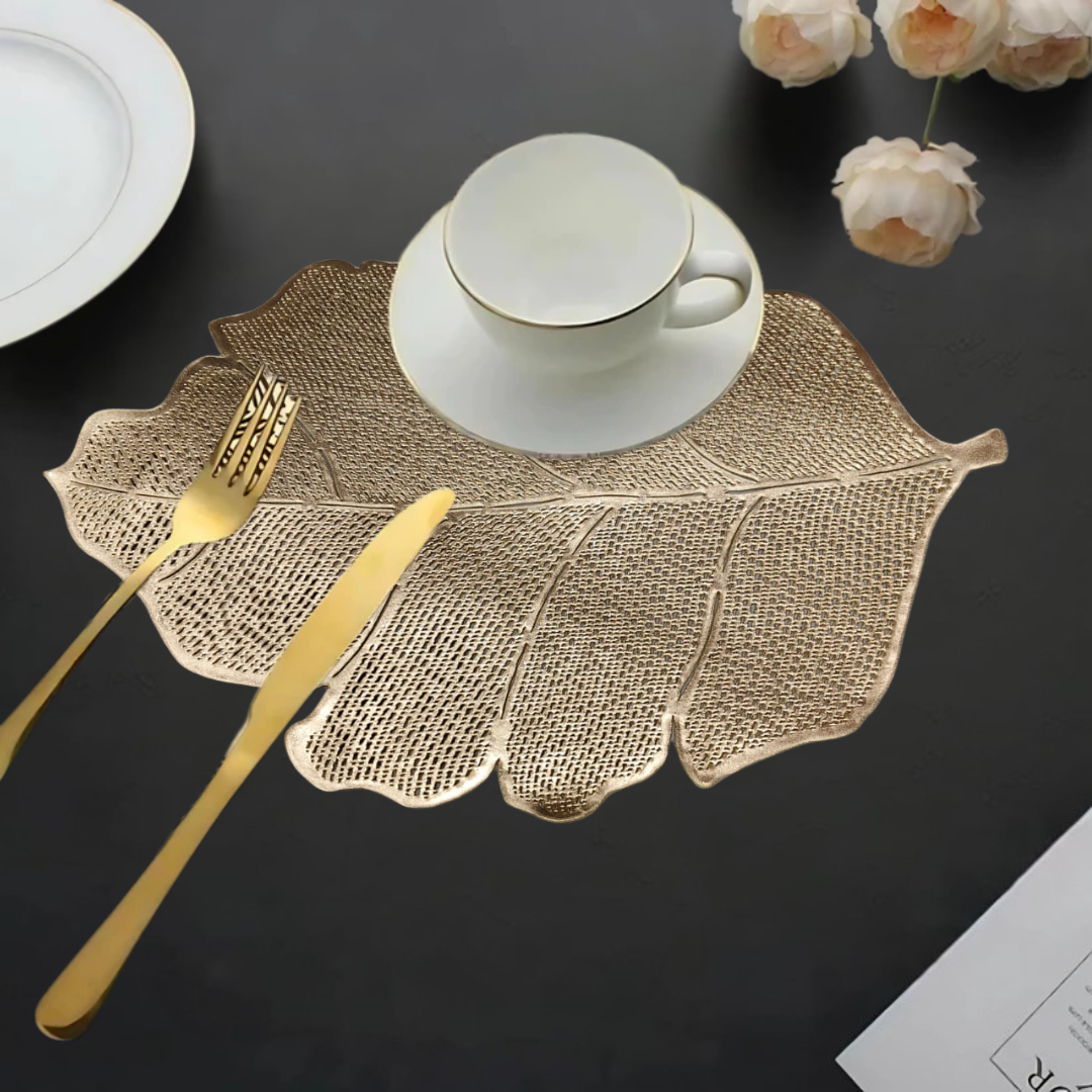 Leaf-shaped gold table mat with a white cup, gold cutlery, and flowers on a dark table.