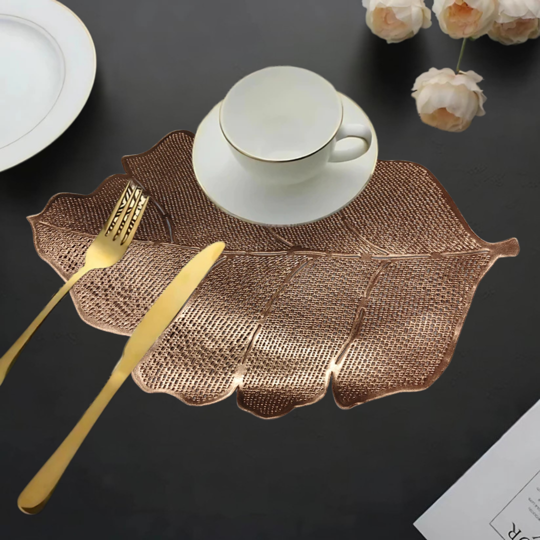 Leaf-shaped copper table mat with a white cup, gold cutlery, and flowers on a dark table.