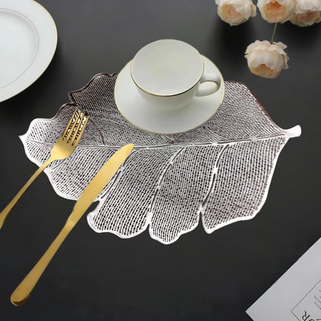 Leaf-shaped silver placemat with a white cup, gold cutlery, and flowers on a dark table.