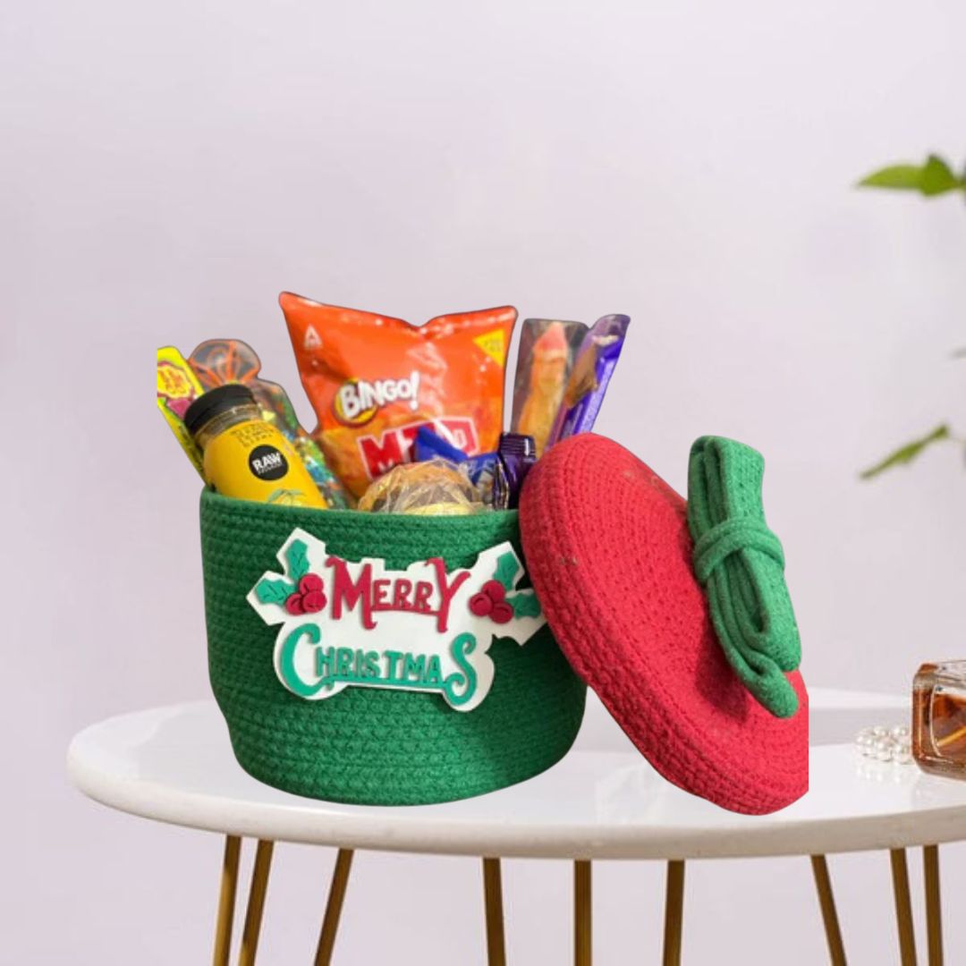 This is a festive green gift basket filled with snacks and decorated with a "Merry Christmas" sign. It comes with a matching red-and-green lid—perfect for holiday gifting!