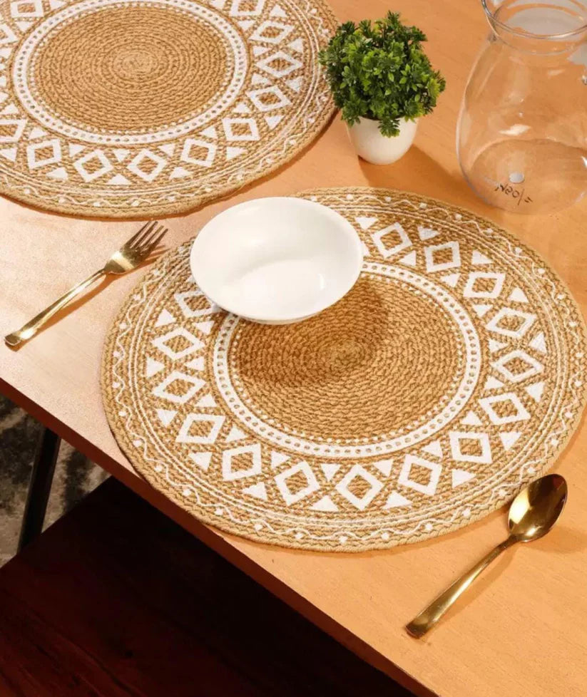Round jute table mat with white geometric patterns, featuring a white bowl and gold cutlery on a wooden table.